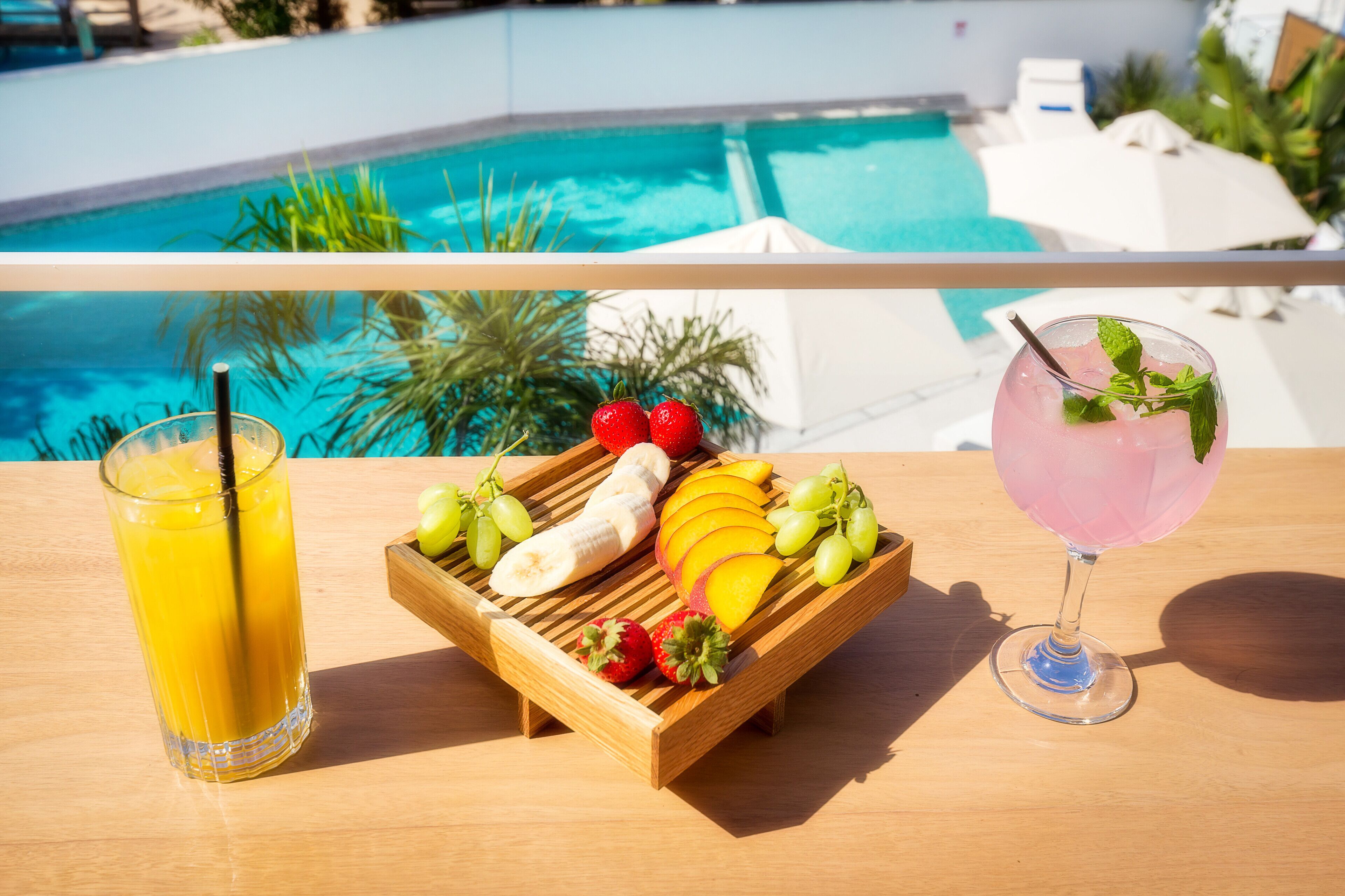 A wooden tray with sliced fruits and a drink on a table by a pool.