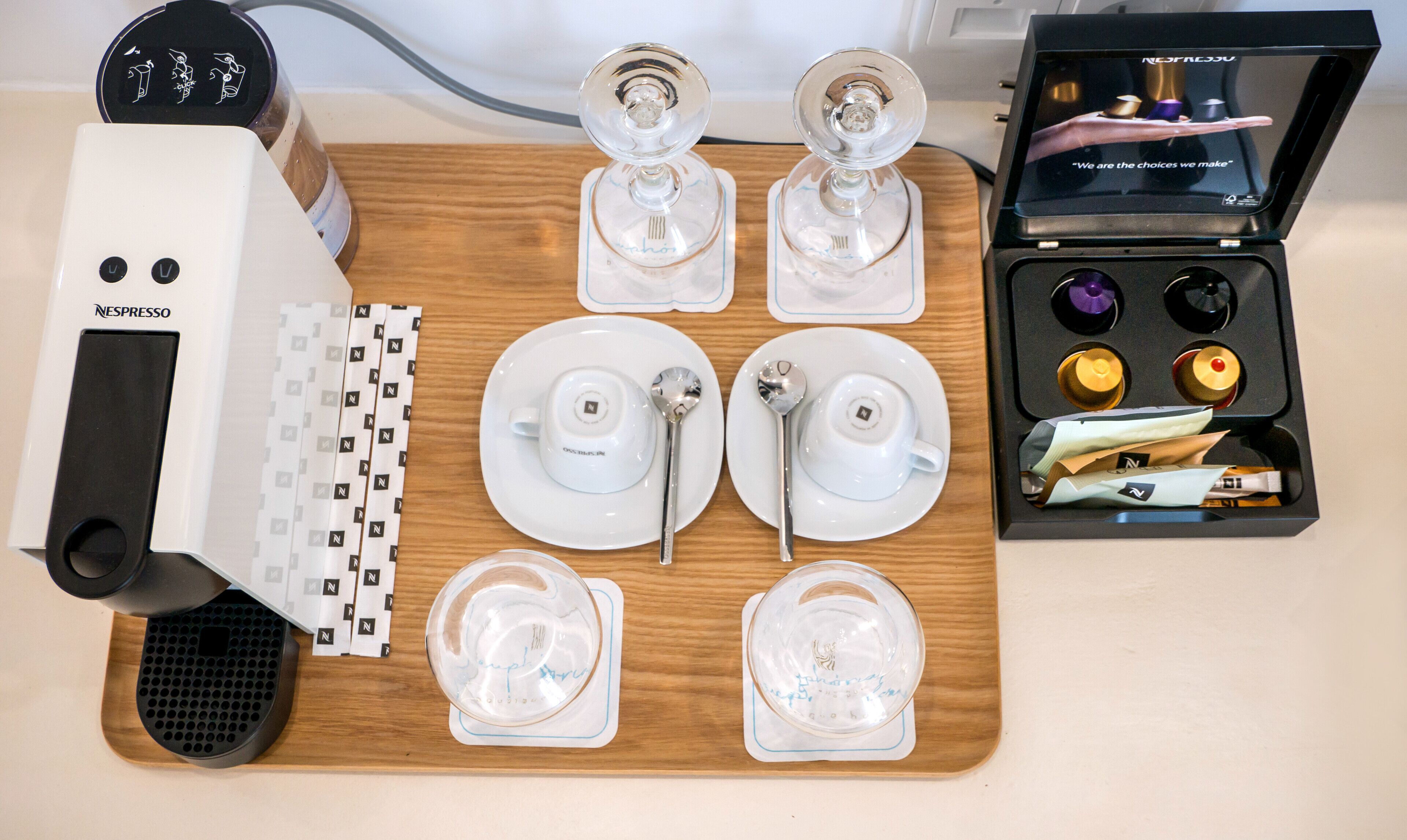 A Nespresso coffee machine, a set of four cups, spoons, and a box of coffee capsules on a wooden tray.