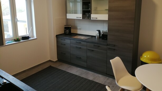 A modern kitchen with dark cabinets, a coffee maker, and a toaster. There is a window with a view outside, a small plant on the windowsill, and a yellow helmet on a table. - Komfortable Ferienwohnung mit Parklplatz (Zossen)