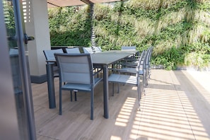 An outdoor patio with a long table and chairs, surrounded by lush greenery.