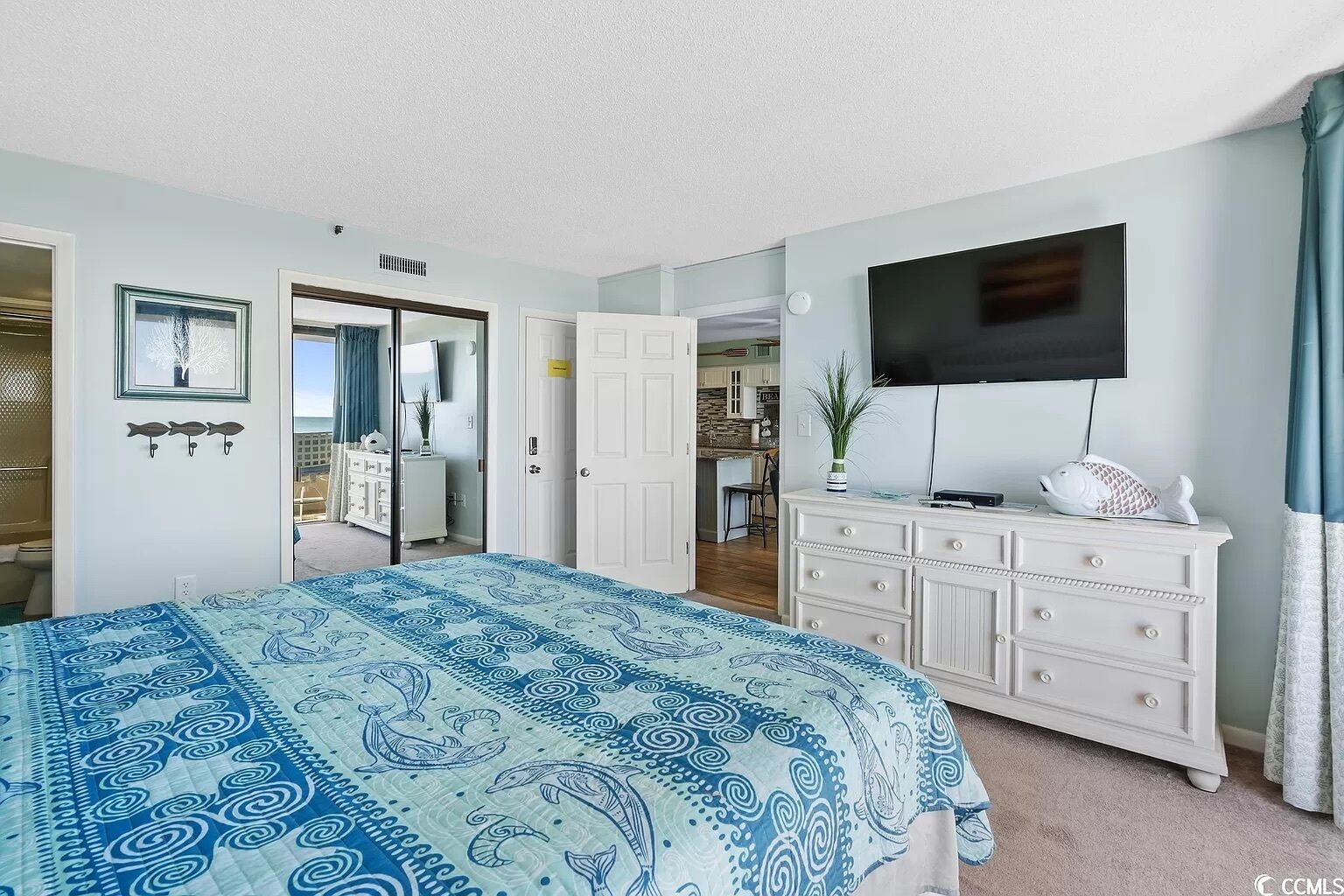 A bedroom with a bed, a dresser, a TV, and a view into the kitchen.