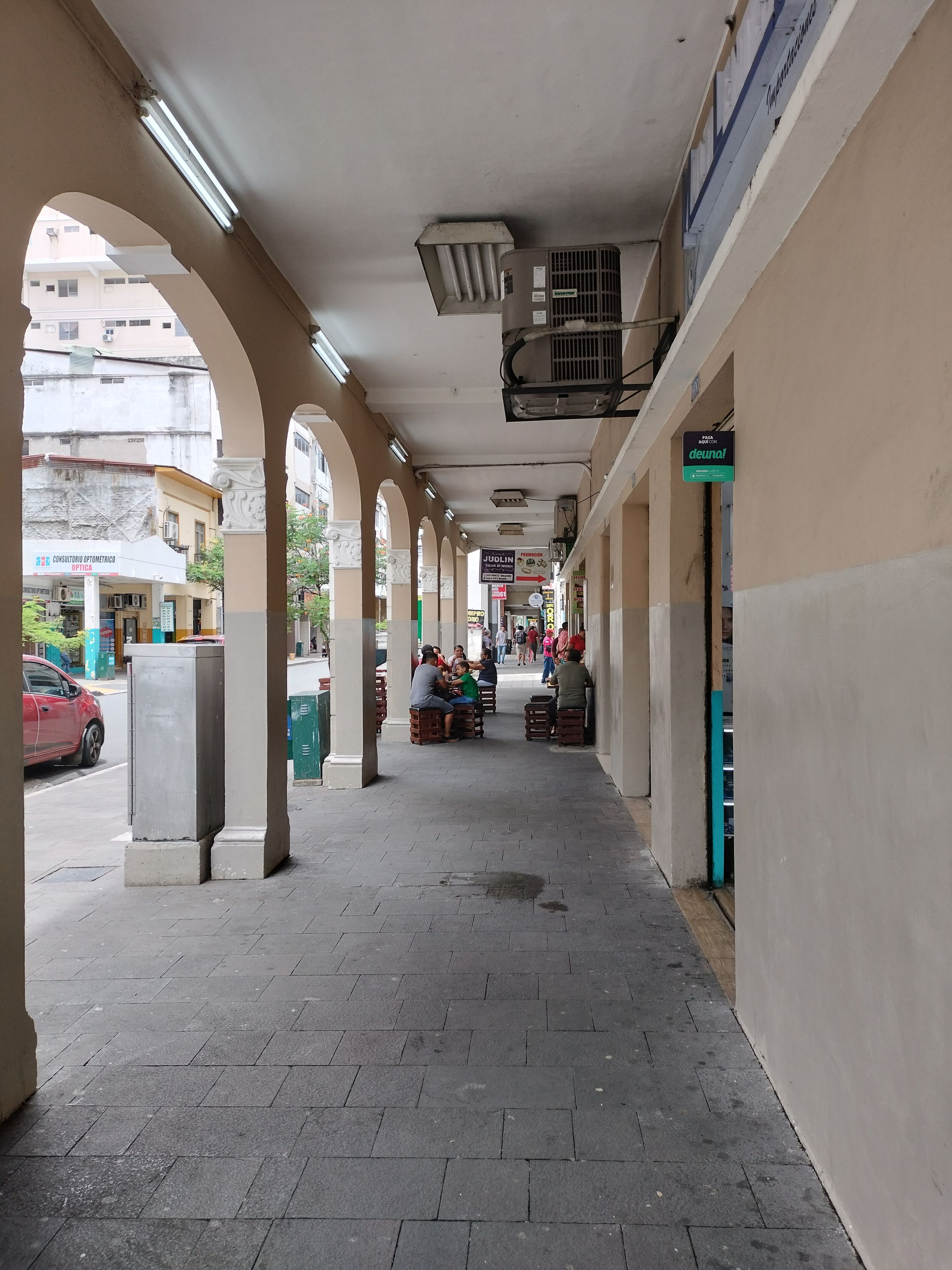 A covered walkway with benches, a storefront, and a red car parked on the side.