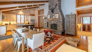 A spacious kitchen with a stone fireplace, wooden beams, and a dining area with a red patterned rug. - Gorgeous Renovated Home With Mirror Lake Access! (Lake Placid)