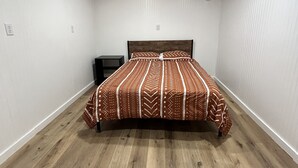 Une chambre avec un plancher en bois, un lit recouvert d’une literie à motifs, une table de chevet et un plafond avec des poutres apparentes et des spots encastrés.