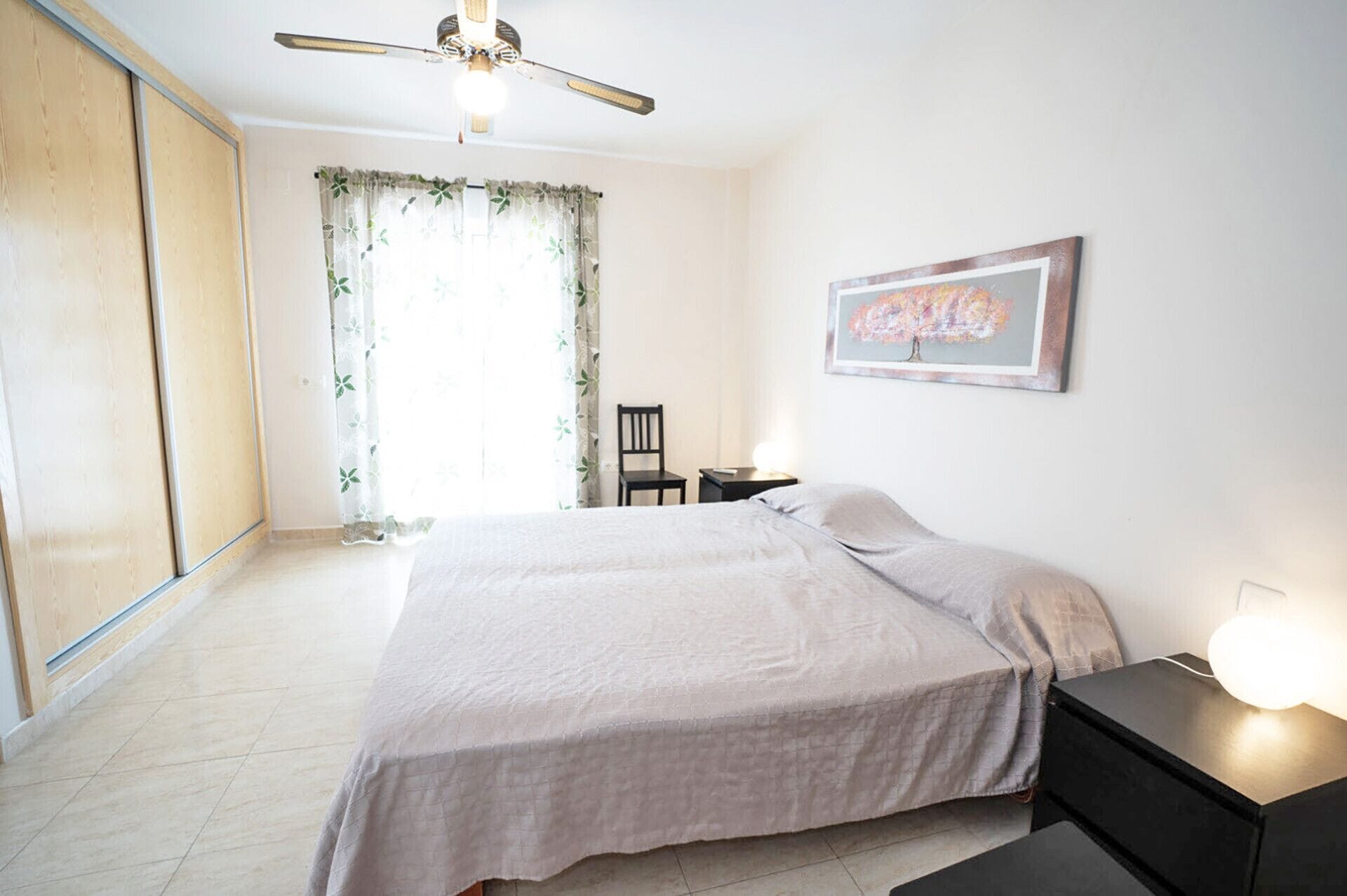 Une chambre à coucher comprenant un lit, une table de chevet, une chaise, un ventilateur de plafond et une fenêtre avec des rideaux.