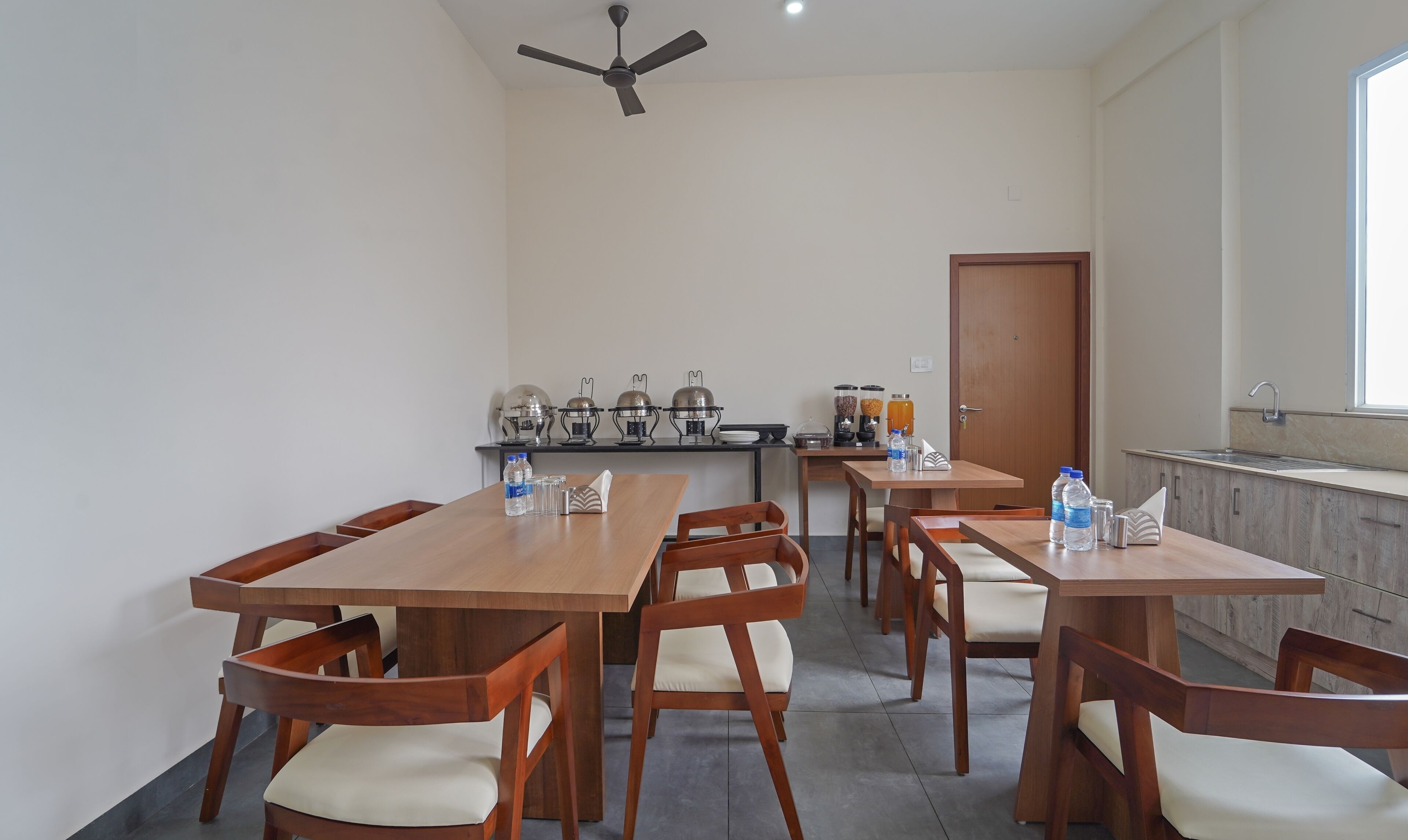 Un comedor con mesas y sillas de madera, un ventilador de techo y una zona de cocina con electrodomésticos y un fregadero.