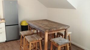 Une petite cuisine avec une table et des tabourets en bois, un réfrigérateur et une poubelle jaune.