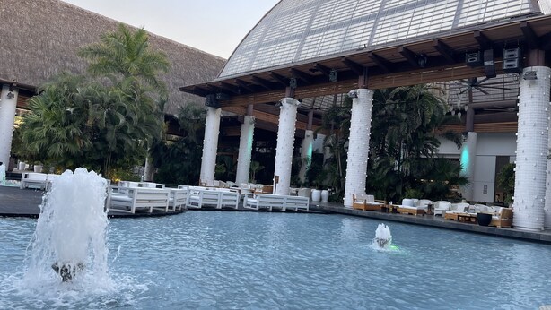 Un área de piscina con una fuente, sillas de descanso y una zona de estar cubierta con techo de cristal.