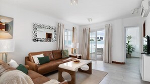 A modern living room with a brown leather sofa, a white coffee table, and a large mirror with a circular frame. There is a window with curtains, a lamp, and a television on a stand.