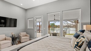 A bedroom with a large bed, two chairs, a small table, and a view of a lake through open doors. - Spectacular, New Waterfront Home.  A Place to Escape and Enjoy Lake Life! (Mabank)