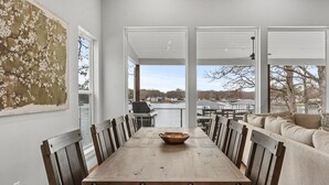 A dining area with a wooden table and chairs, a large painting on the wall, and a view of the outdoors through large windows. - Spectacular, New Waterfront Home.  A Place to Escape and Enjoy Lake Life! (Mabank)