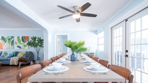 Une salle à manger avec une longue table en bois, dressée pour six personnes, entourée de chaises en bois dotées de coussins bleu clair. Un ventilateur de plafond est visible au-dessus de la table, et la pièce est dotée de grandes fenêtres sur un côté.