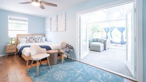 Une chambre avec un lit, une chaise, une petite table, un ventilateur de plafond et une vue sur un espace extérieur aménagé, accessible par une porte-fenêtre coulissante.