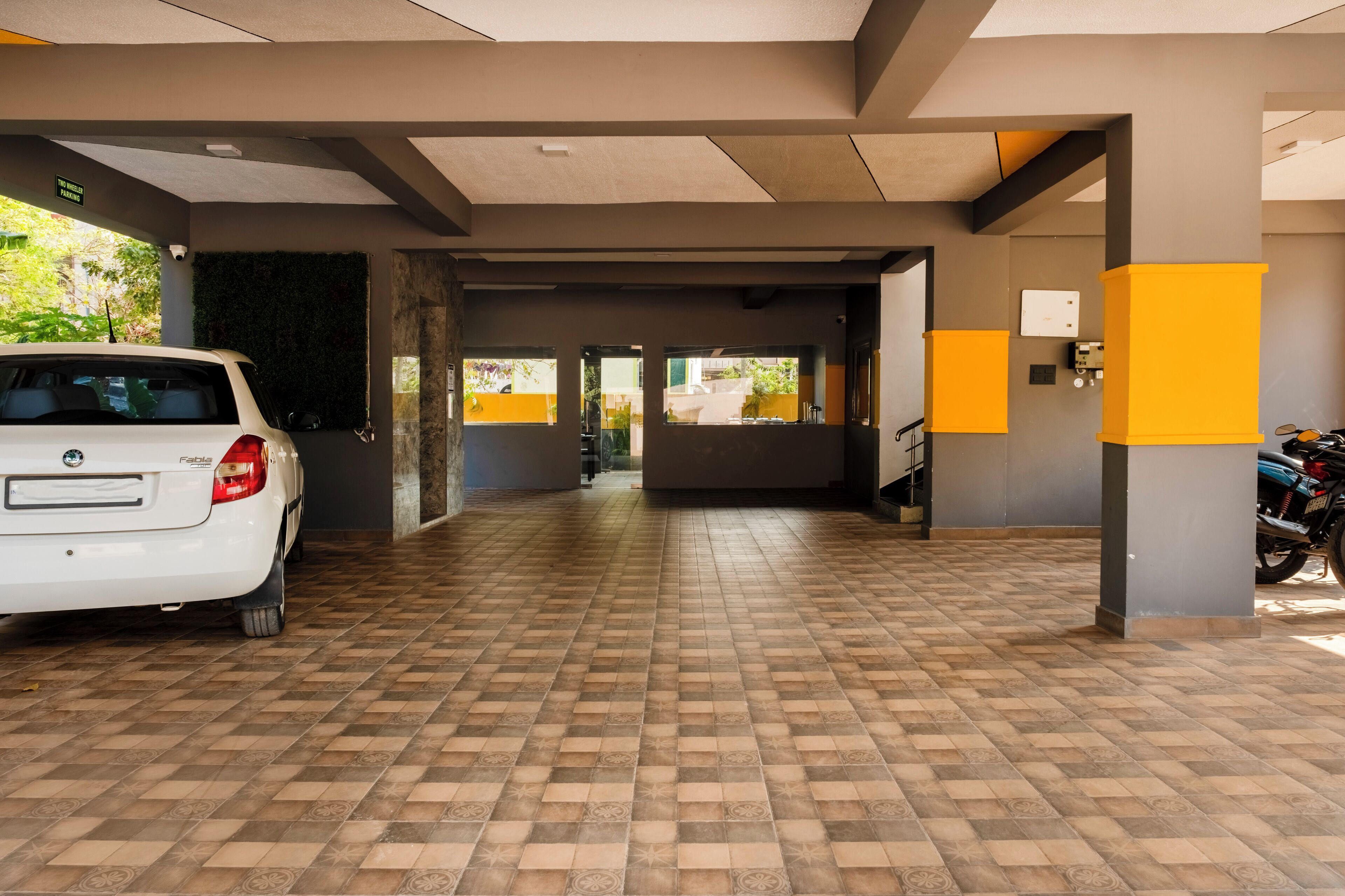 A covered parking area with a white car parked on the left, a motorcycle on the right, and a view of the outside through the windows.