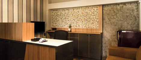A compact hotel room with a kitchenette, a wooden desk, a sofa, and a television.