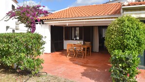 A covered patio with a tiled roof, a wooden table and chairs, and a small garden area with shrubs and a flowering plant.