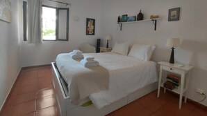 A neatly made bed with towels, a bedside table with a lamp, a nightstand, and a window with curtains.