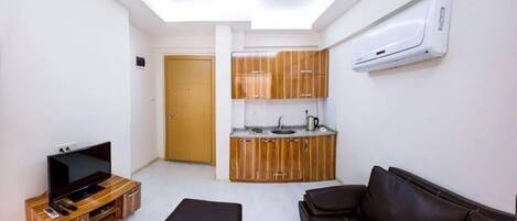 A compact living space with a kitchenette, a television on a wooden stand, a brown leather sofa, and a ceiling light fixture.