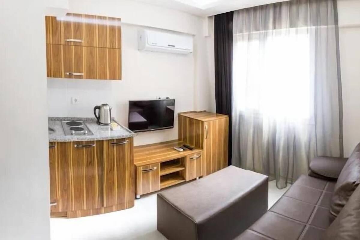 A modern living room with a kitchen area, a flat-screen TV, wooden cabinets, a sofa, and a large window with curtains.