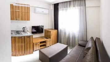 A modern living room with a kitchen area, a flat-screen TV, wooden cabinets, a sofa, and a large window with curtains.