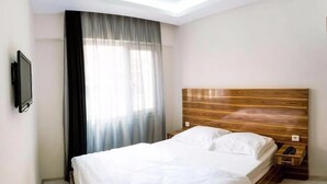 Hotelkamer met een bed, een houten hoofdeinde, nachtkastje, een aan de muur bevestigde televisie en een raam met dunne gordijnen.