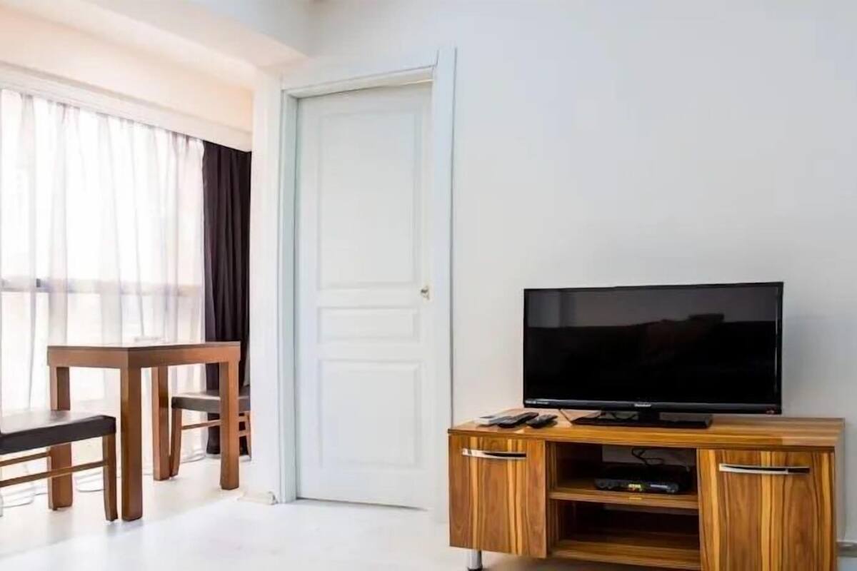 A hotel room with a wooden TV stand, a flat-screen TV, a small table with chairs, and a large window with sheer curtains.