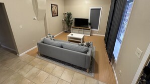 Uma sala de estar com um sofá cinzento, uma mesa de centro, uma televisão em um móvel, uma planta e uma janela com cortinas.