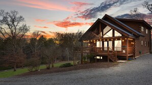 A log cabin with a porch and large windows, surrounded by trees and a gravel driveway.