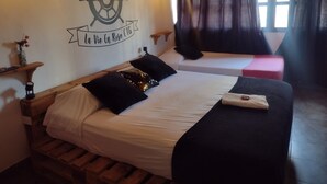 A room with two beds, a wooden headboard, a wall with a ship's wheel and text, a window with curtains, and a small shelf with a vase.