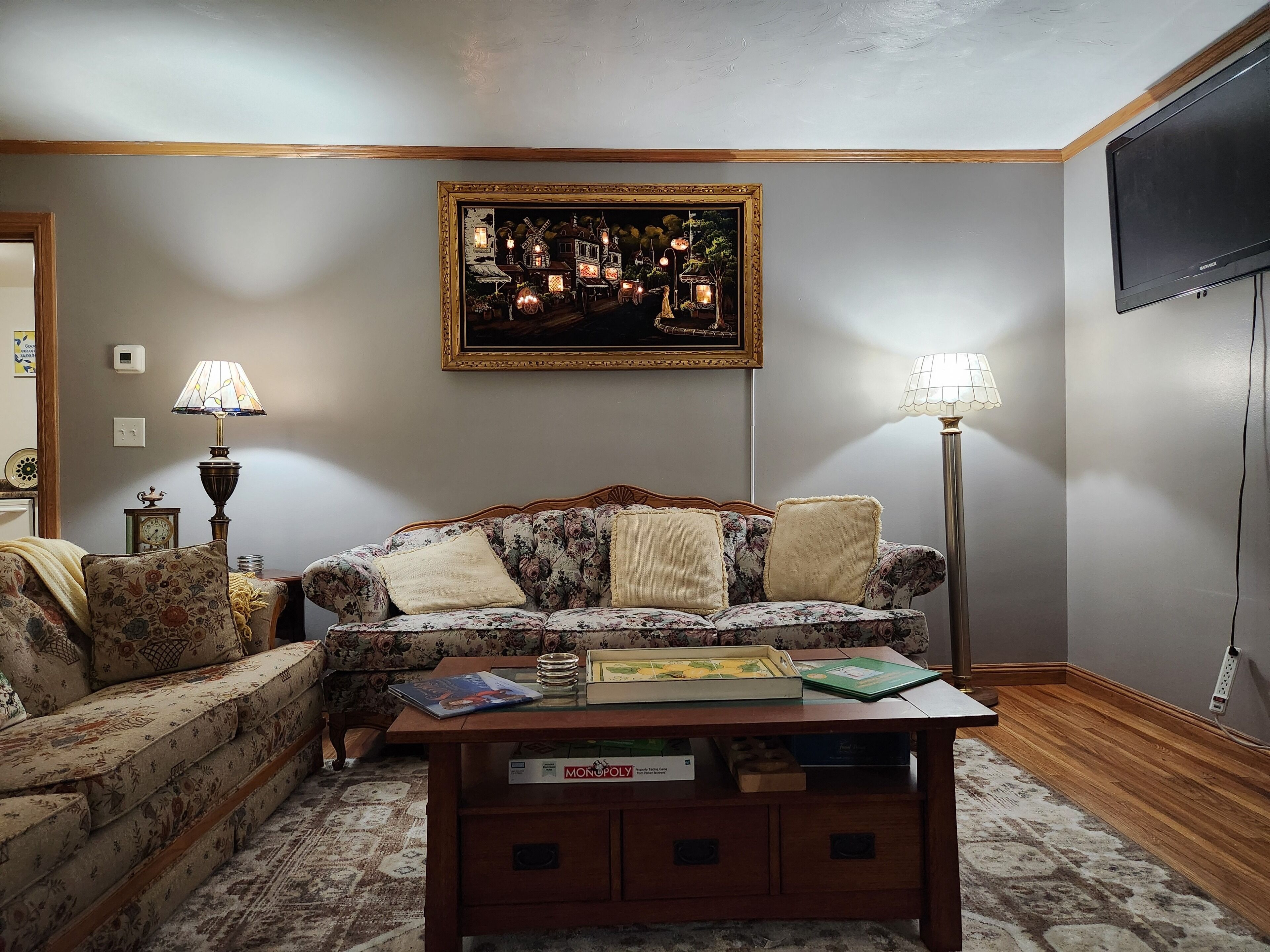 A living room with a floral sofa, a coffee table with board games, a framed painting, and a floor lamp.