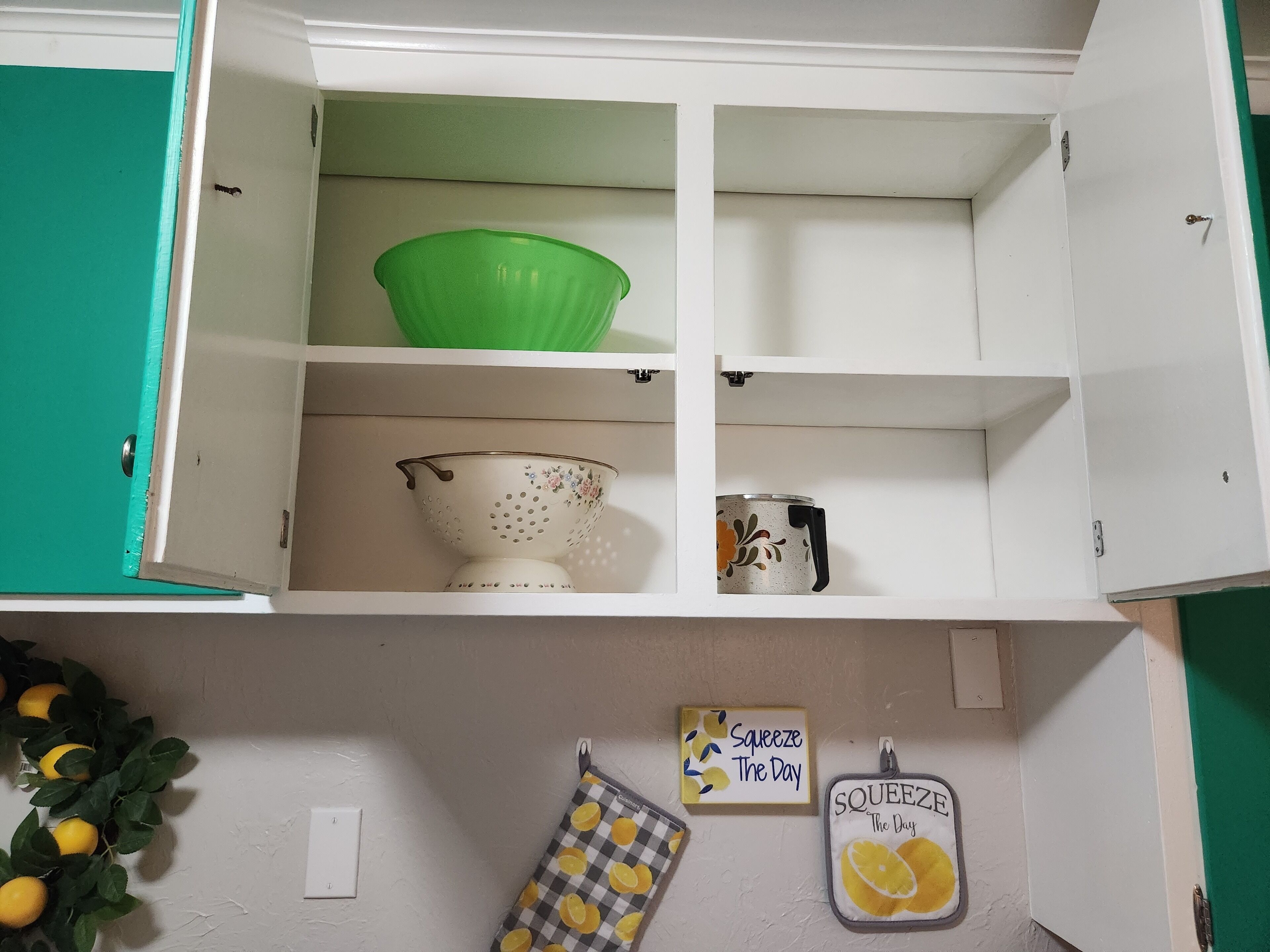 A kitchen cabinet with a green bowl, a floral sieve, and a mug. There are two kitchen towels hanging on the wall.