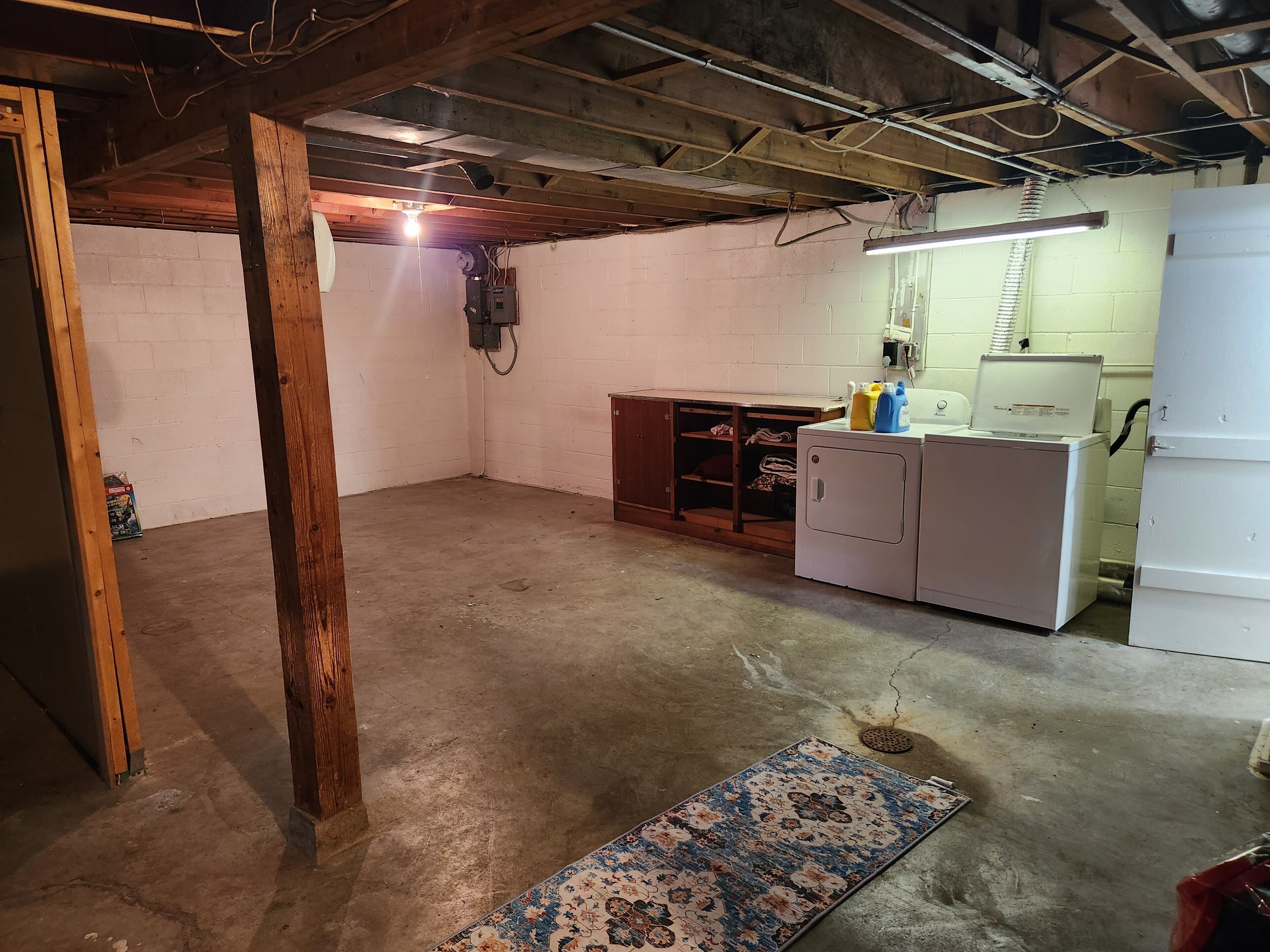 A basement with exposed wooden beams, a washing machine, and a utility room with a light fixture.
