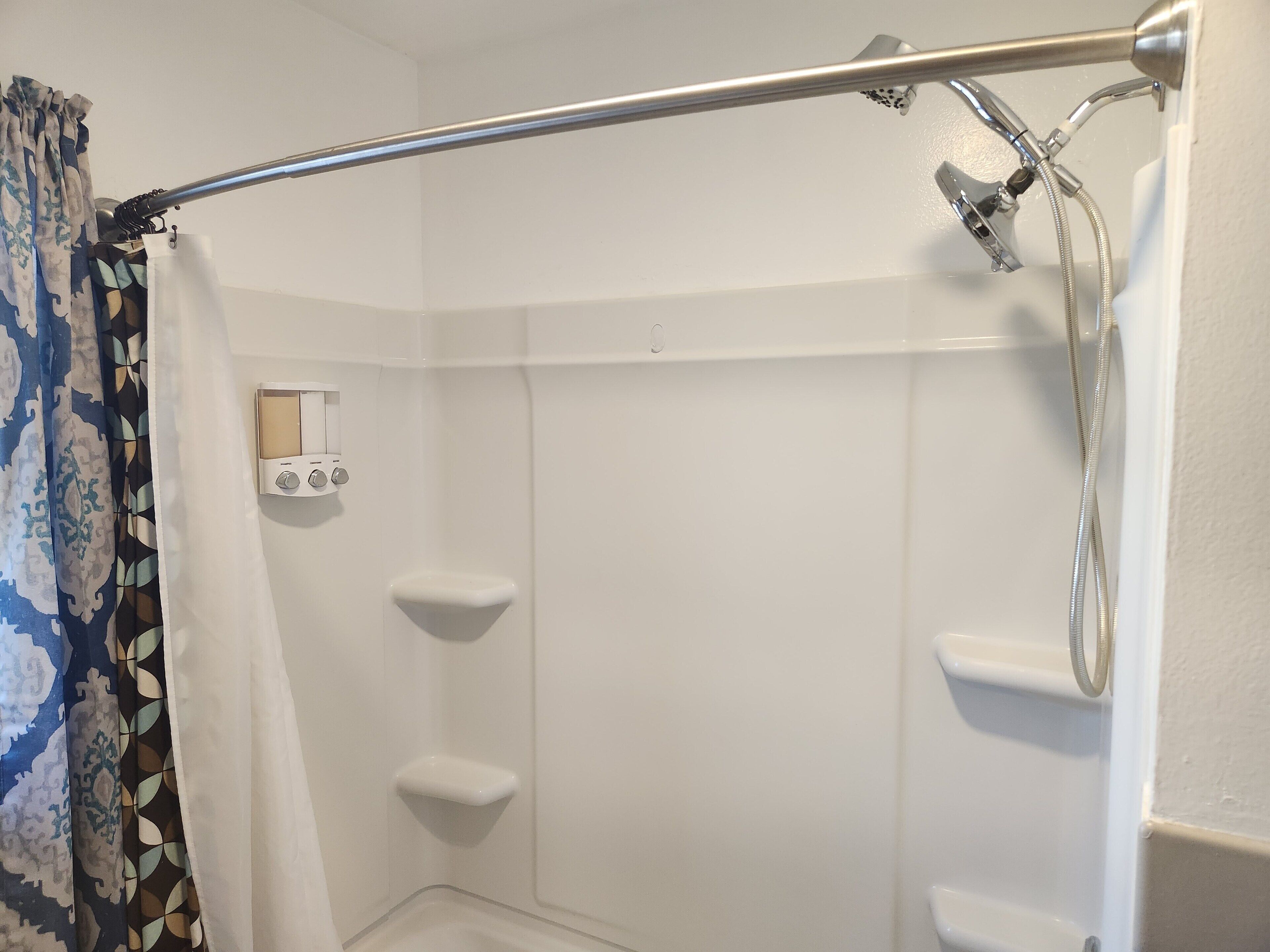 Una ducha con cabezal de ducha manual, panel de control y pared con azulejos blancos.