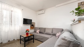 Una sala de estar con un sofá gris, una mesita con una maceta roja, un televisor de pantalla plana y un equipo de aire acondicionado.