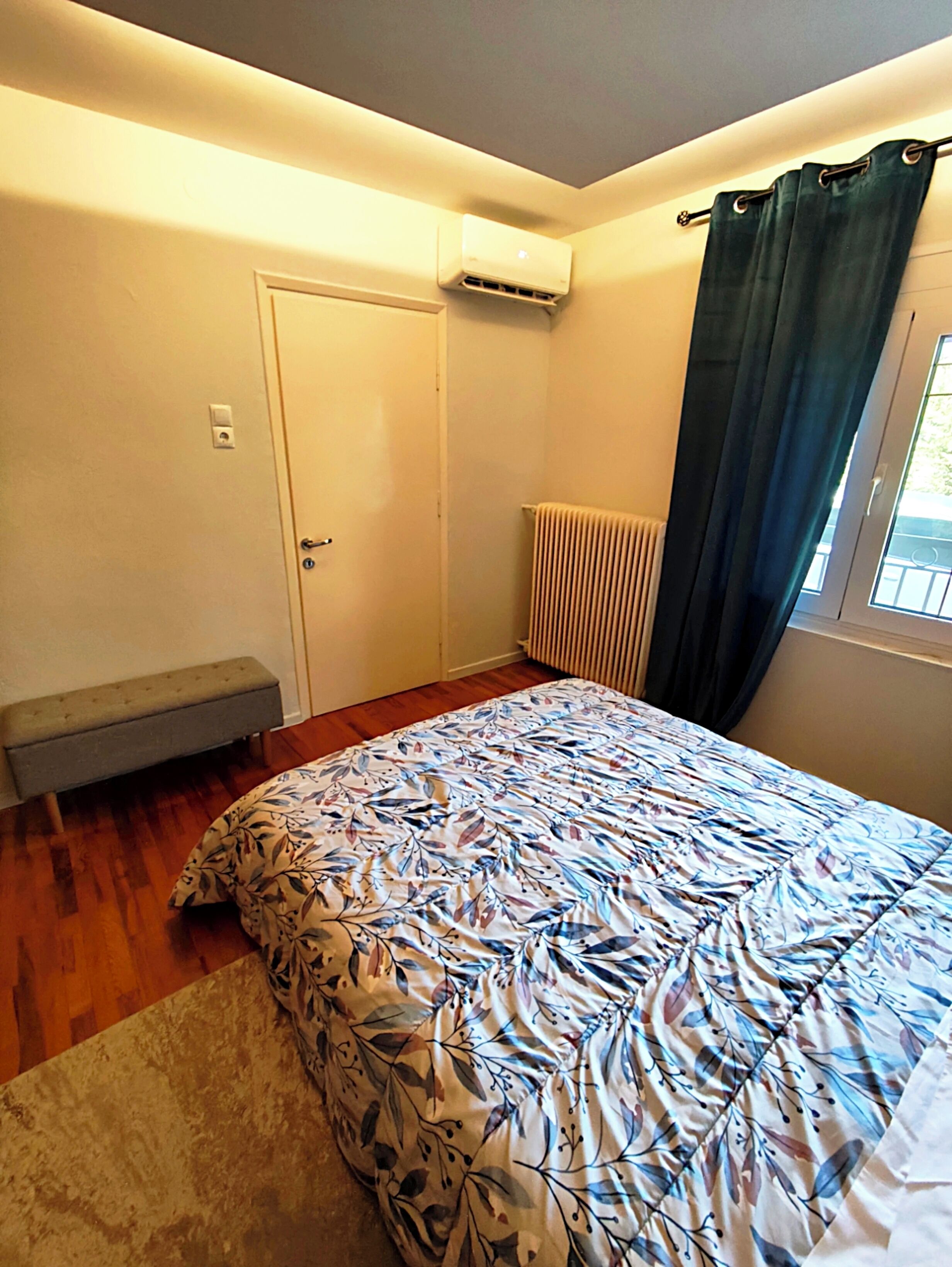 A bedroom with a bed, a bench, a radiator, an air conditioning unit, and a window with curtains.