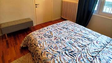 A bedroom with a bed, a bench, a radiator, an air conditioning unit, and a window with curtains.
