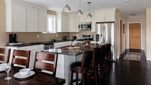 A spacious kitchen with white cabinets, a granite countertop, and stainless steel appliances. There are three pendant lights, a large island with bar stools, and a dining table set for two. - River & Charm: 2 Homes, on 10 Acres Near Clayton (Clayton)