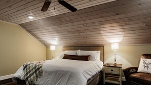 A bedroom with a wooden ceiling, a bed with white bedding, two bedside lamps, and a brown leather chair. - River & Charm: 2 Homes, on 10 Acres Near Clayton (Clayton)