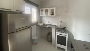 Eine kompakte Küche mit einem Edelstahlkühlschrank, einem Gasherd und einem kleinen Backofen. Ein Fenster mit Jalousien, ein Spülbecken und ein Mülleimer sind vorhanden.