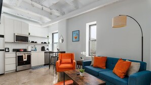 Una cocina moderna con armarios blancos, un comedor con sofá azul y cojines naranjas, y un ventanal que proporciona luz natural.