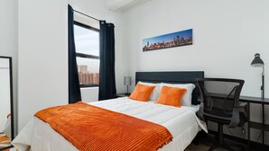 Habitación de hotel con cama, escritorio, silla, lámpara, ventana con cortinas y un cuadro de paisaje urbano en la pared.