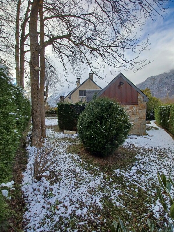 A house with a stone foundation, a red barn-like structure, and a snow-covered yard with trees and bushes.