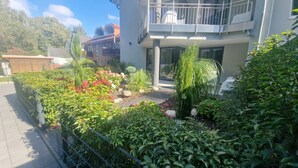 Een woongebouw met een balkon, een tuin met diverse planten en bloemen, en een pad dat naar de ingang leidt.