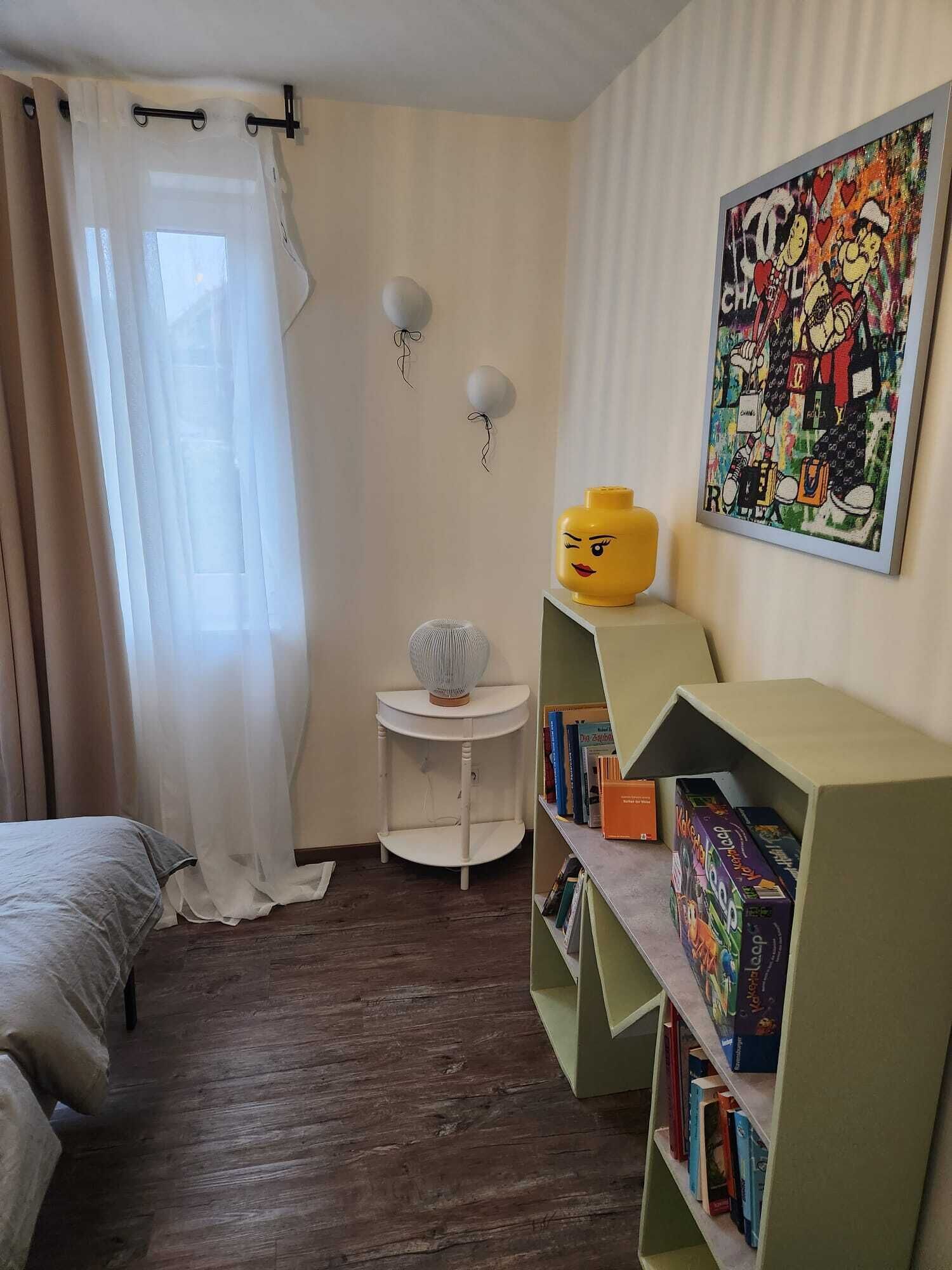 Ein Schlafzimmer mit einem Bett, einem kleinen weißen Nachttisch, einem Bücherregal, einem Fenster mit durchsichtigen Vorhängen und einem Wandbild.