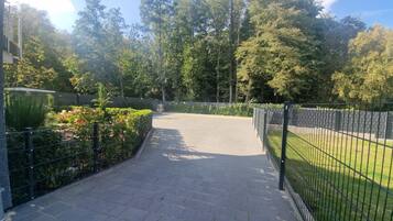 A paved pathway leading to a gated entrance surrounded by lush greenery and a metal fence.
