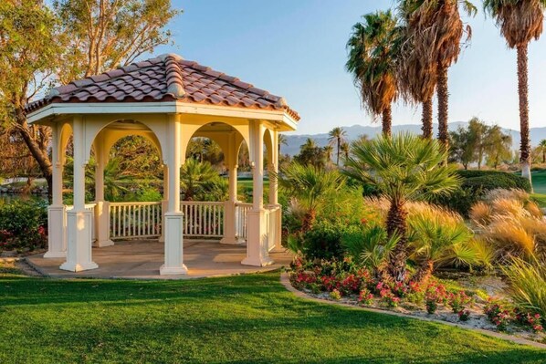 Situated in a lush garden, this gazebo features a tiled roof and white columns, surrounded by a white fence and palm trees.