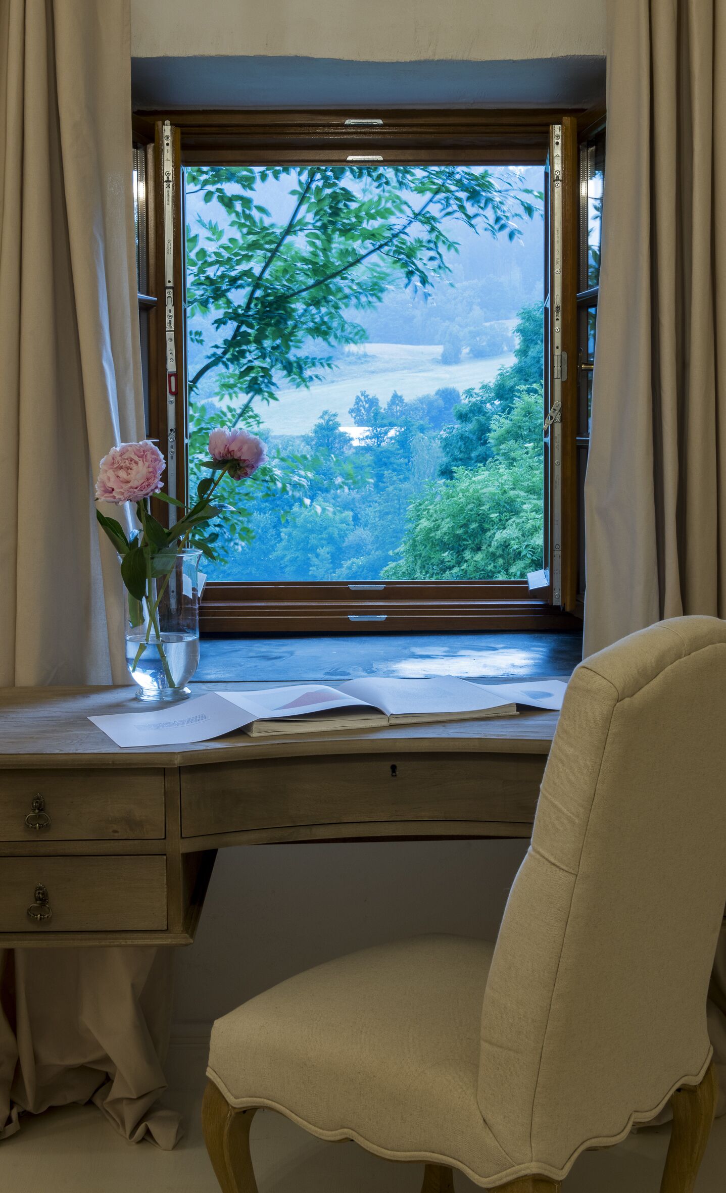A room with a desk, a chair, a window with curtains, and a vase with flowers.