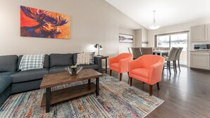 A modern living room with a sofa, coffee table, and two armchairs. A colorful abstract painting is on the wall. - 5-Br/3-Ba | Private Pool & Hot Tub | Pool Table + Home Theatre (Hinton)
