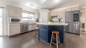 A modern kitchen with white cabinetry, stainless steel appliances, a large island with stools, and a window with a view of trees. - 5-Br/3-Ba | Private Pool & Hot Tub | Pool Table + Home Theatre (Hinton)