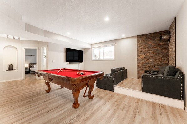 Amplia sala de estar con mesa de billar, televisor de pantalla plana montado en la pared, pared decorativa de piedra y un rincón de descanso acogedor con sofá y sillón.